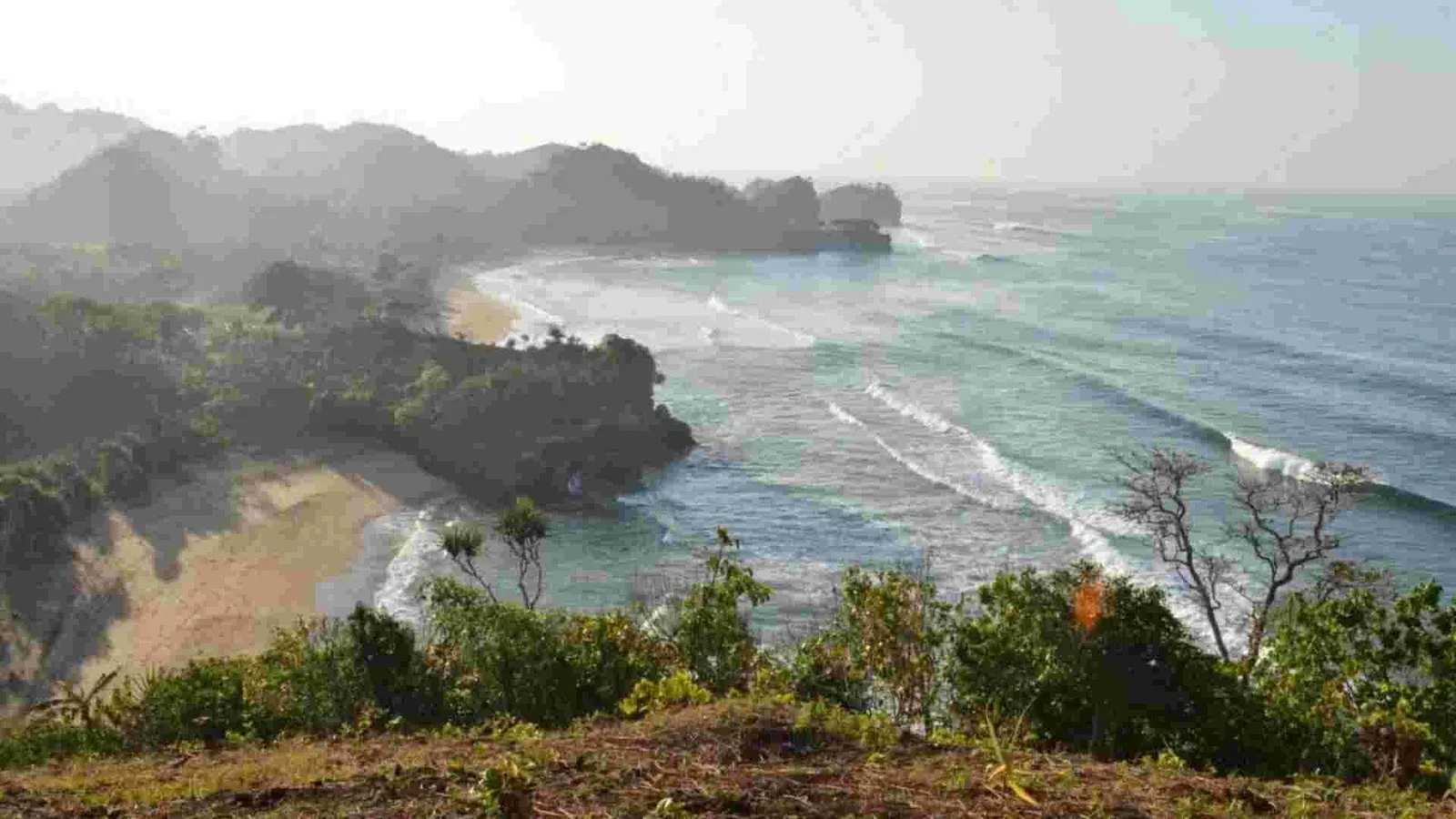Panorama teluk Pantai Nganteb dari ketinggian dengan ombak putih dan tanjung hijau